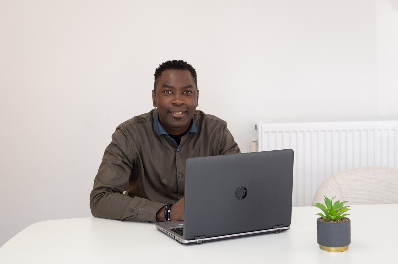 Een man zit aan een witte tafel met een open HP laptop voor zich en een kleine potplant ernaast.