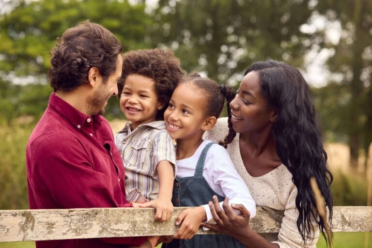 Vier mensen van gemengde bevolkingsgroepen staan buiten bij een houten hek en glimlachen naar elkaar. Twee volwassenen houden twee kinderen vast, met bomen en groen op de achtergrond, die de diversiteit van Afrikaanse landen vastleggen.